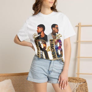May include: A white short-sleeved t-shirt featuring a graphic print of two men, Usher and Chris Brown, with the text RAYMOND and BROWN integrated into large letters spelling R&B. The design includes gold-colored script signatures and small photo collages.