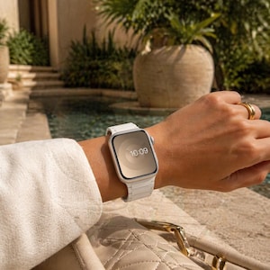 Op de afbeelding: Een witte smartwatch met een rechthoekige wijzerplaat die de tijd 10:09 weergeeft. Het horloge heeft een witte band en wordt om de pols gedragen. Een gouden ring is zichtbaar op de hand. De achtergrond toont een zwembad en groen.