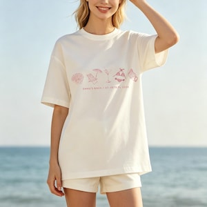 May include: Off-white t-shirt with a pink beach-themed graphic and the text "EMMA'S BACH / ST. PETE, FL 2026". The shirt is paired with off-white shorts. The background shows a sunny beach and ocean.