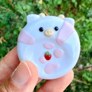 May include: A round, light blue glass trinket dish shaped like a cow face with small horns, pink patches, black eyes, a beige snout, and a small red strawberry detail in the center. The surface is smooth, glossy, and reflective.