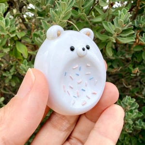 May include: A small, smooth, white glass bear figurine with a rounded shape. The bear features two small ears with gold-colored centers, two black bead-like eyes, and a central belly area decorated with scattered pink and light blue sprinkles.
