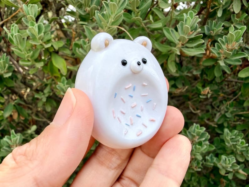 May include: A small, smooth, white glass bear figurine with a rounded shape. The bear features two small ears with gold-colored centers, two black bead-like eyes, and a central belly area decorated with scattered pink and light blue sprinkles.