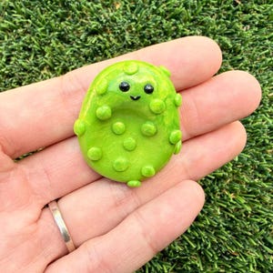 May include: A handmade, lime green clay charm shaped like a smiling pickle. The charm has small, round bumps and black eyes. The charm is held in a hand against a background of green grass.