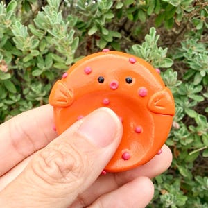 May include: A bright orange, circular pufferfish figurine with a smooth, glossy finish. The creature features two small black eyes, a tiny circular mouth, and several small pink raised dots scattered across its surface, along with two side fins.