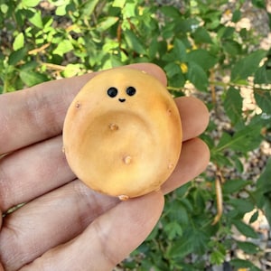 May include: A small, round, tan-colored decorative item with a smiling face and small black eyes. The item has a few small, raised bumps and is held in a person's hand against a backdrop of green foliage.