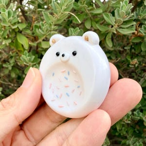 May include: A small, round, white ceramic bear figurine with two small ears, two black dot eyes, and a tiny nose. The center of the bear features a recessed circular area decorated with small, scattered pink and light blue sprinkles.