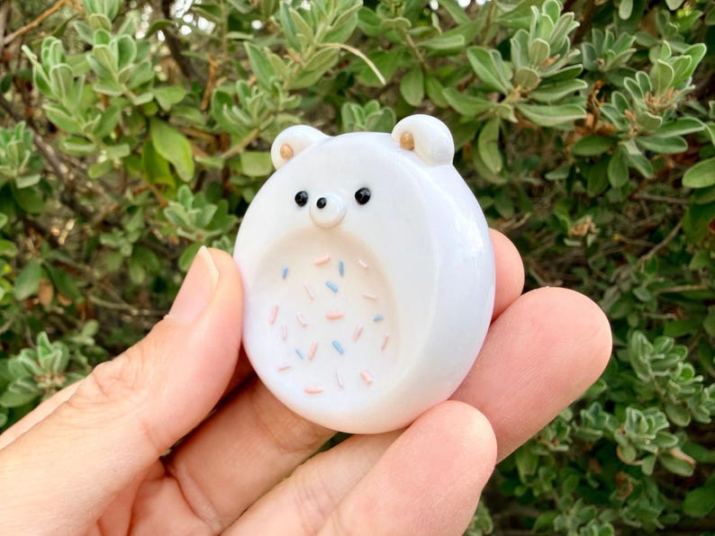 May include: A small, round, white ceramic bear figurine with two small ears, two black dot eyes, and a tiny nose. The center of the bear features a recessed circular area decorated with small, scattered pink and light blue sprinkles.