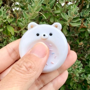 May include: A round, white, smooth, glossy, bear-shaped fidget stone with two small ears, two black eyes, a small black nose, and a recessed center area decorated with tiny, colorful, scattered sprinkles on the surface of the object.
