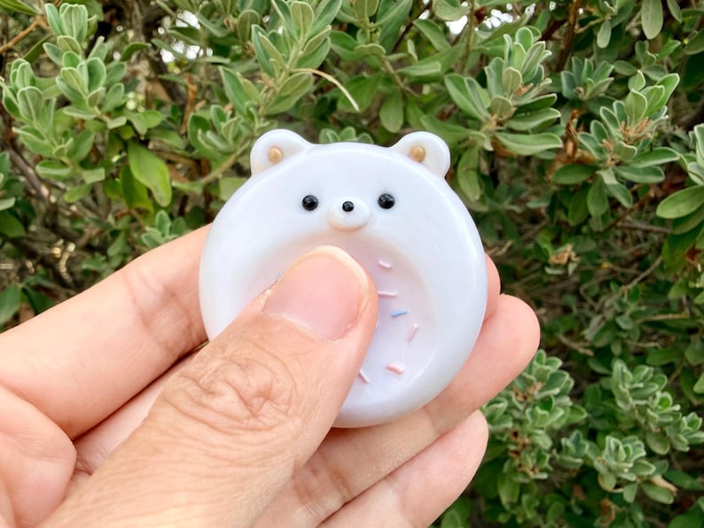 May include: A round, white, smooth, glossy, bear-shaped fidget stone with two small ears, two black eyes, a small black nose, and a recessed center area decorated with tiny, colorful, scattered sprinkles on the surface of the object.