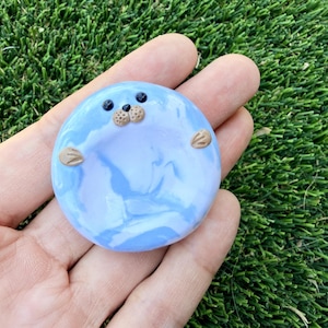 May include: A round, blue and white marbled clay seal figurine with black eyes, a tan nose, and tan flipper-like arms. The seal is held in a hand against a green background.