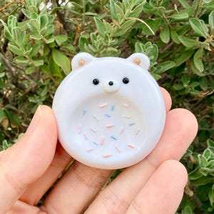 May include: A circular white glass trinket dish shaped like a bear face with two small ears, black bead eyes, and a small nose. The center of the dish features a pattern of small, scattered pink and light blue sprinkles embedded in the smooth surface.