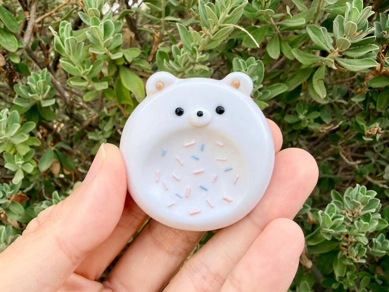 May include: A circular white glass trinket dish shaped like a bear face with two small ears, black bead eyes, and a small nose. The center of the dish features a pattern of small, scattered pink and light blue sprinkles embedded in the smooth surface.