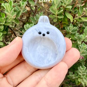 May include: A small, circular, light blue marbled ceramic dish featuring a stylized rabbit head at the top with two small ears and three tiny black dots representing the eyes and nose. The dish has a smooth, glossy finish and a concave center.