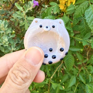 May include: A small, round, light lavender glass trinket dish featuring a stylized cat face with two small ears, two black dot eyes, a white nose, and several black polka dots scattered across the lower portion of the smooth, glossy surface.