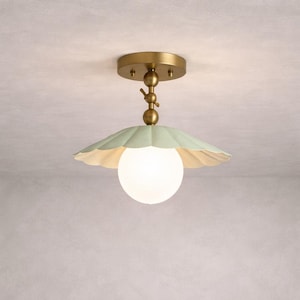 Puede incluir: Una lámpara de techo con acabado de latón y una pantalla verde pálido con volantes. La pantalla rodea una luz blanca esférica. El accesorio está diseñado con una estética decorativa y vintage.