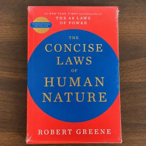 May include: A hardcover book titled "The Concise Laws of Human Nature" by Robert Greene. The book has a red cover with a blue circle in the center. The text is in gold and white. The book is shrink-wrapped.