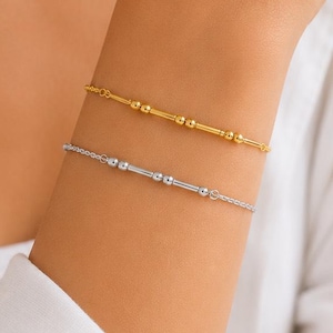 May include: Two delicate bracelets, one gold and one silver, are displayed on an arm. The gold bracelet features small, evenly spaced beads, while the silver one has a similar design with rectangular and round elements. The bracelets are on a white background.