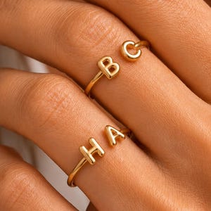 May include: Two gold-toned rings featuring uppercase letters. The top ring displays the letters B and C, while the bottom ring displays the letters H and A. Each ring has a thin, polished band and a smooth, rounded finish on the metallic letters.