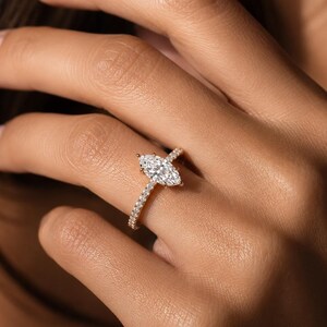May include: A gold ring featuring a large marquise-cut center diamond held by prongs, with a thin band encrusted with small round pavé diamonds extending along the sides. The metal has a warm yellow tone, and the stones are clear and faceted.