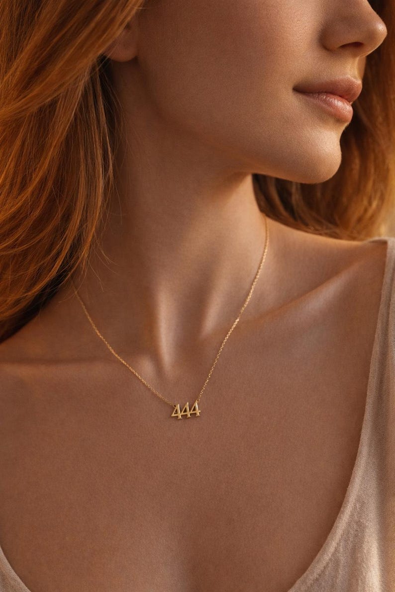 May include: A gold-toned necklace featuring a delicate chain and a pendant shaped as the number 444. The pendant is centered on the chain, which rests against the neck. The metal has a polished finish that reflects light, creating a bright appearance.