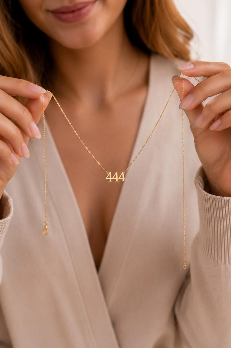 May include: A gold-toned necklace featuring a pendant with the number 444 in a serif font. The pendant is suspended from a delicate, thin gold-toned chain with a small lobster claw clasp visible at the end of the chain link structure.