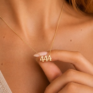 May include: A gold-toned necklace featuring a pendant shaped as the number 444. The pendant is suspended from a delicate, thin gold-toned chain. The design consists of three identical numerals in a serif font, connected to the chain at the top.