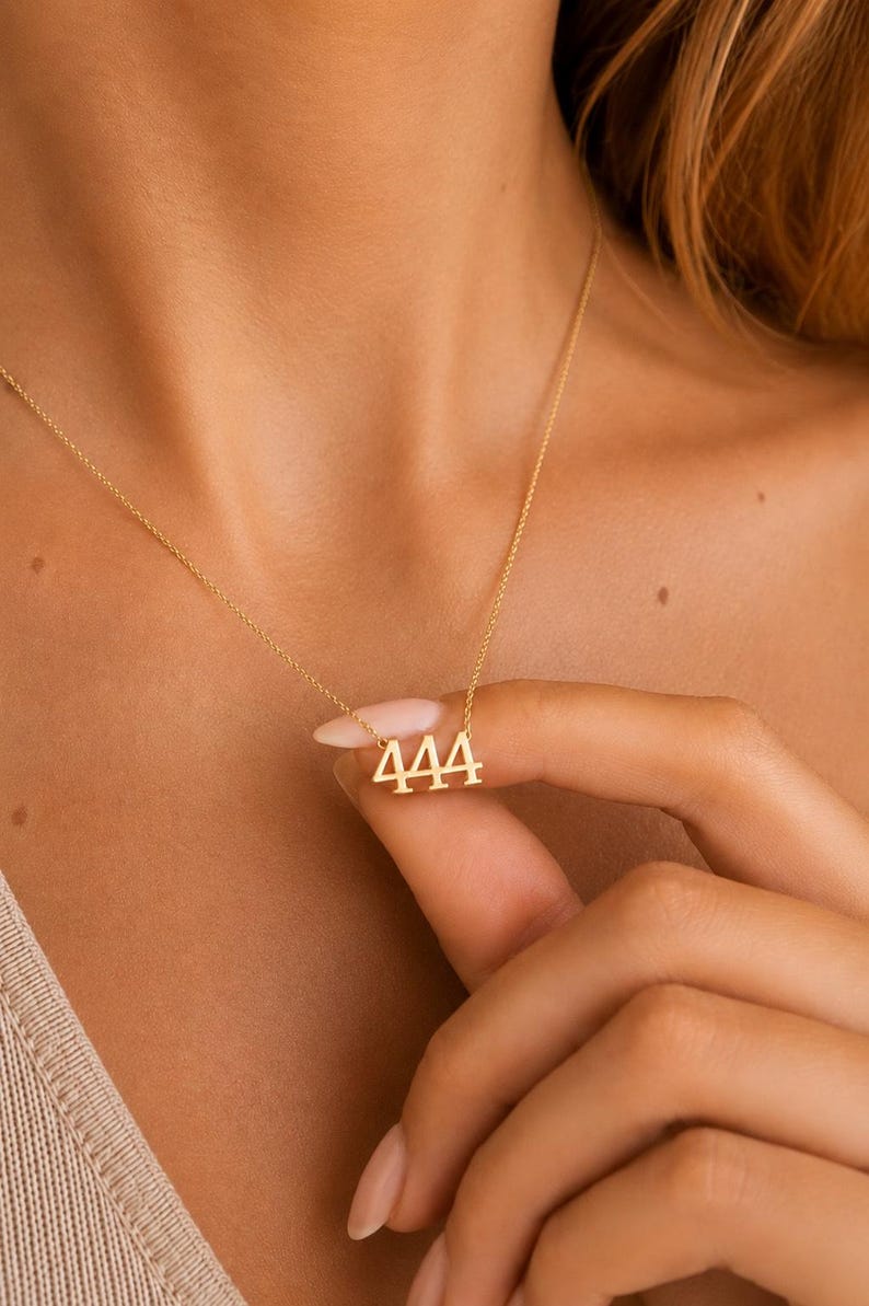 May include: A gold-toned necklace featuring a pendant shaped as the number 444. The pendant is suspended from a delicate, thin gold-toned chain. The design consists of three identical numerals in a serif font, connected to the chain at the top.