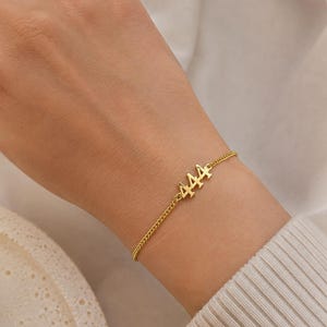 May include: A gold-colored bracelet with the number "444" charm is displayed on a wrist. The bracelet has a delicate chain design. The background is a soft, neutral color, highlighting the jewelry.