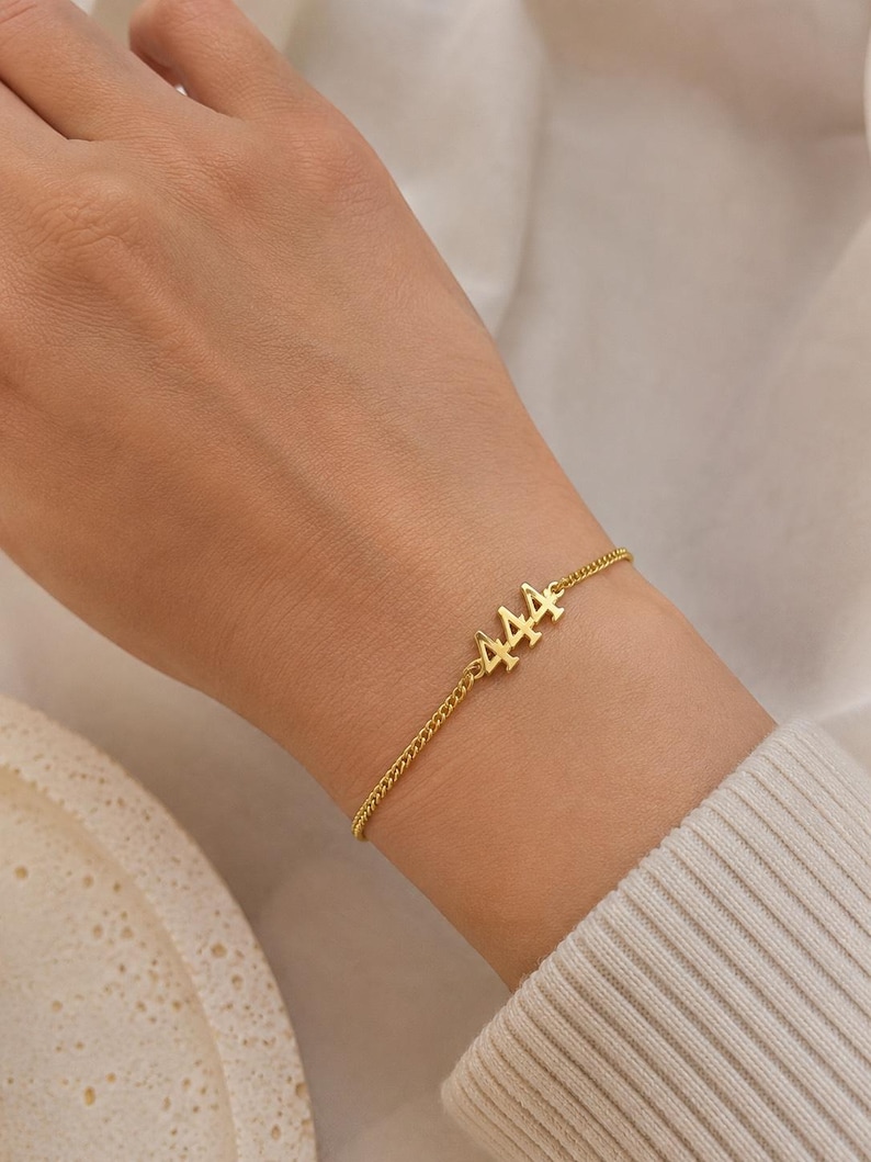 May include: A gold-colored bracelet with the number "444" charm is displayed on a wrist. The bracelet has a delicate chain design. The background is a soft, neutral color, highlighting the jewelry.