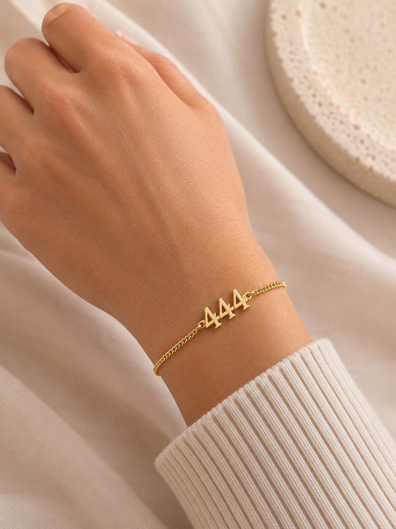 May include: A gold-colored bracelet featuring the number "444" in a delicate font. The bracelet is on a fine chain and is worn on a wrist, against a white sweater. The bracelet is a simple, elegant piece of jewelry.