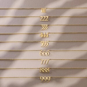 May include: A collection of gold-colored necklaces, each featuring a different number pendant: 111, 222, 333, 444, 555, 666, 777, 888, and 999. The necklaces are displayed on a soft, neutral-toned surface.