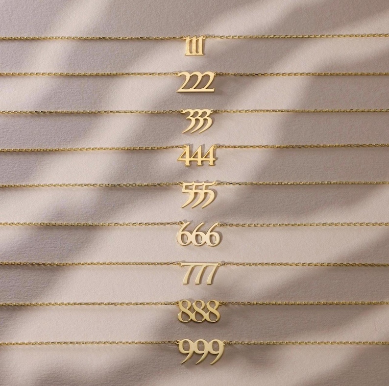 May include: A collection of gold-colored necklaces, each featuring a different number pendant: 111, 222, 333, 444, 555, 666, 777, 888, and 999. The necklaces are displayed on a soft, neutral-toned surface.
