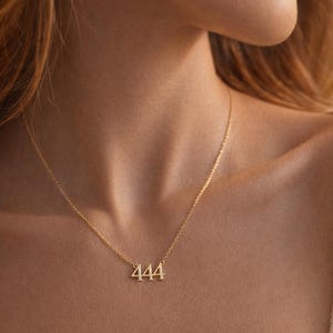 May include: A gold-toned necklace featuring a pendant shaped as the number 444. The pendant is suspended from a delicate, thin gold-toned chain that rests around the neck. The metal has a polished, reflective finish that catches the light.