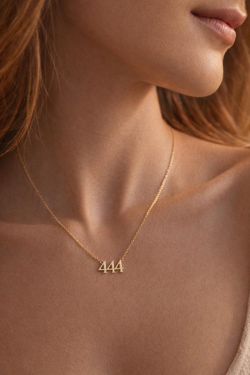 May include: A gold-toned necklace featuring a pendant shaped as the number 444. The pendant is suspended from a delicate, thin gold-toned chain that rests around the neck. The metal has a polished, reflective finish that catches the light.