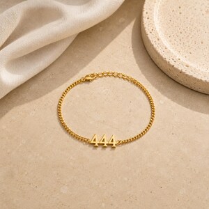 May include: Gold-colored chain bracelet featuring the number 444. The bracelet has a delicate chain design and a lobster clasp closure. The bracelet is displayed on a neutral-toned surface.