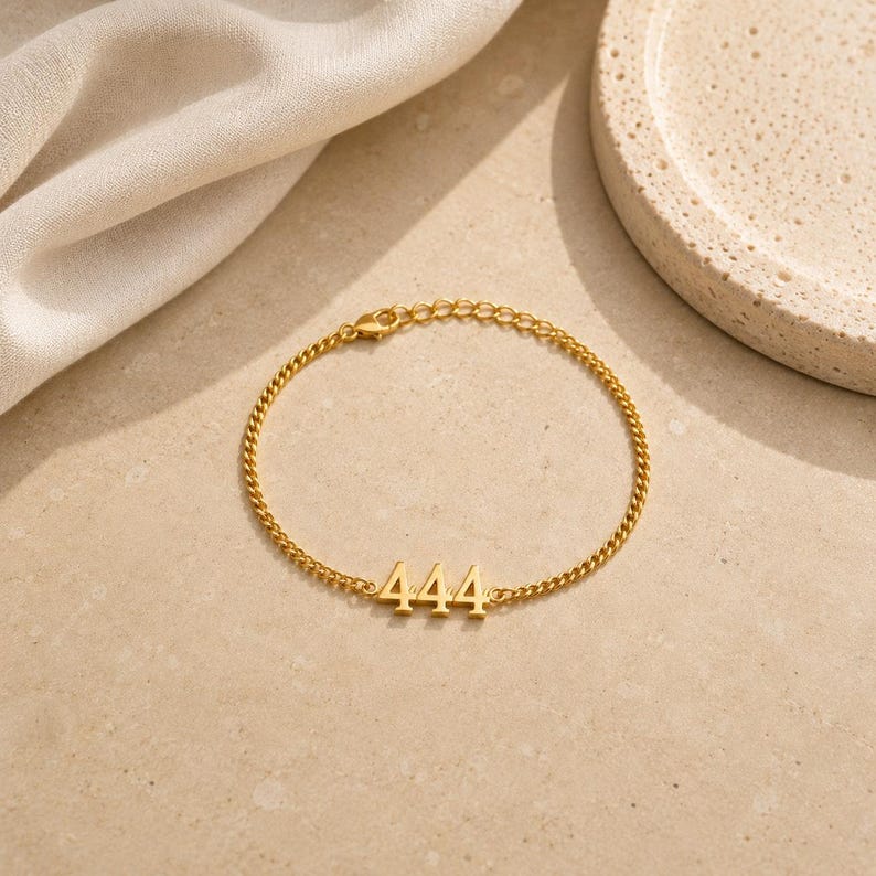 May include: Gold-colored chain bracelet featuring the number 444. The bracelet has a delicate chain design and a lobster clasp closure. The bracelet is displayed on a neutral-toned surface.