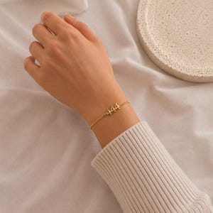 May include: A gold-colored bracelet with the number "444" is worn on a wrist. The bracelet is delicate, with a thin chain. The wrist is covered by a cream-colored sweater. A white, textured tray is in the background.