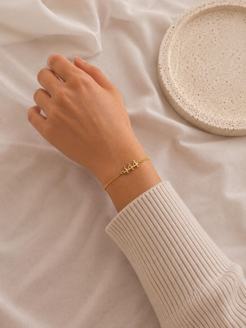May include: A gold-colored bracelet with the number "444" is worn on a wrist. The bracelet is delicate, with a thin chain. The wrist is covered by a cream-colored sweater. A white, textured tray is in the background.