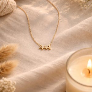 May include: A gold-toned necklace featuring a pendant with the number 444 in a serif font. The pendant is attached to a delicate, thin gold-toned chain. The necklace is displayed against a soft, light-colored fabric background.