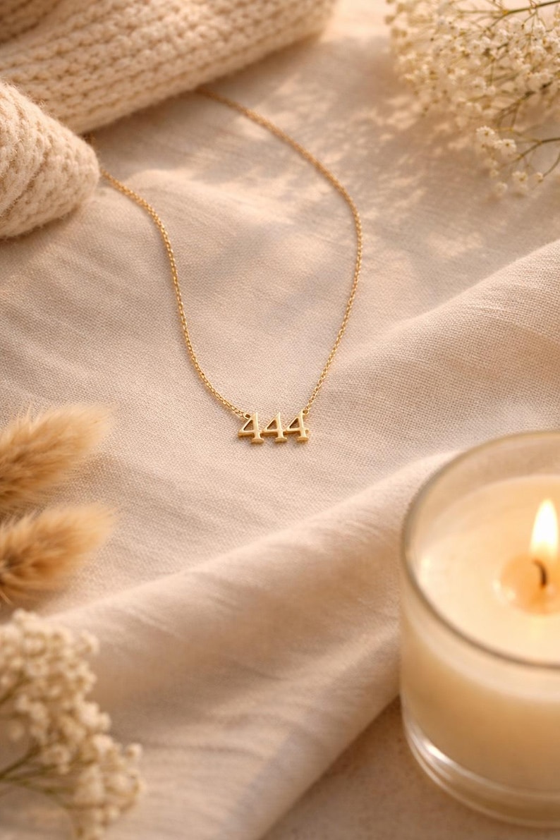May include: A gold-toned necklace featuring a pendant with the number 444 in a serif font. The pendant is attached to a delicate, thin gold-toned chain. The necklace is displayed against a soft, light-colored fabric background.