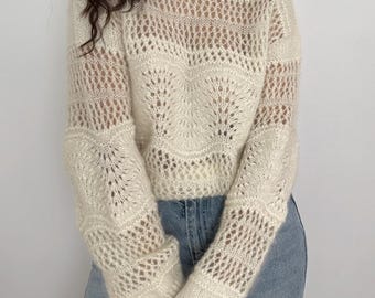 May include: Cream-colored, open-knit sweater with a round neckline and long sleeves. The sweater features intricate patterns, including openwork designs on the sleeves and a central floral motif. The model is wearing blue jeans.