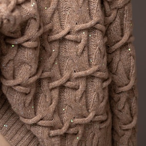 May include: A tan cable-knit sweater with a textured design. The sweater features a complex pattern of interwoven cables and is embellished with small, scattered sequins. The sleeves and body of the sweater are visible.