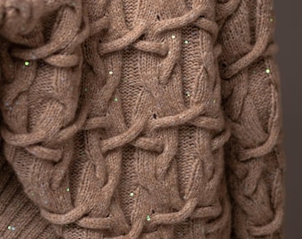May include: A tan cable-knit sweater with a textured design. The sweater features a complex pattern of interwoven cables and is embellished with small, scattered sequins. The sleeves and body of the sweater are visible.