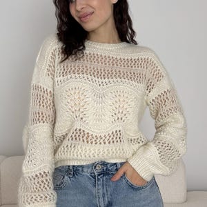 Puede incluir: Suéter de punto abierto color crema, con cuello redondo y mangas largas. El suéter presenta un diseño texturizado y estampado en el pecho y las mangas. La modelo lleva vaqueros de mezclilla azul claro. Una adición elegante a cualquier armario.