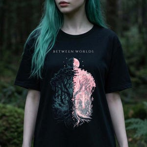 May include: Black t-shirt featuring the text "BETWEEN WORLDS" above a split graphic. One side shows a dark forest, the other a pink forest with a full moon. The design includes trees and a digital glitch effect.