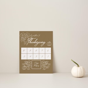 Può includere: Un cartello rettangolare marrone con disegni a linee bianche di fiori e zucche, il testo "Countdown To Thanksgiving" e una griglia con i numeri da 10 a 1. Una piccola zucca bianca è posizionata a destra del cartello su una superficie.