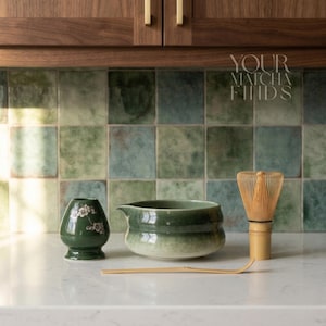 May include: A matcha tea set comprising a dark green ceramic chawan bowl, a matching tea caddy with floral details, a bamboo whisk, and a bamboo scoop. The set is arranged on a white countertop with a green and blue tiled backsplash. The text "YOUR MATCHA FINDS" is visible.