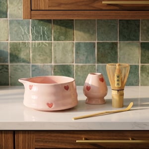 Puede incluir: Un cuenco de matcha de cerámica rosa, un pequeño jarrón de cerámica rosa, un batidor de bambú y una cuchara de bambú están dispuestos sobre una encimera blanca. Cada pieza de cerámica rosa está decorada con formas de corazón rojo. El texto "YOUR MATCHA FINDS" es visible.