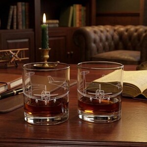 May include: Two clear whiskey glasses with a white airplane design, filled with amber liquid. The glasses sit on a wooden surface, next to a pen, a book, and a small model airplane. A lit green candle is in the background.