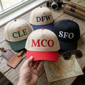 May include: Four baseball caps with embroidered airport codes: CLE, DFW, MCO, and SFO. The caps have different color combinations, including tan, black, and red. Accessories include a vintage camera, sunglasses, and a map.